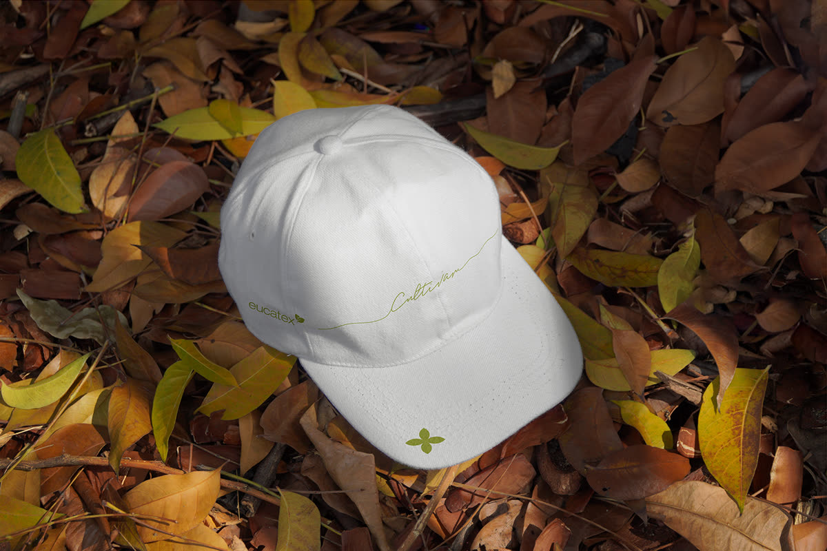 Boné branco com a assinatura Cultivar em verde na copa e o símbolo de folhas na aba, deitado sobre folhas secas.