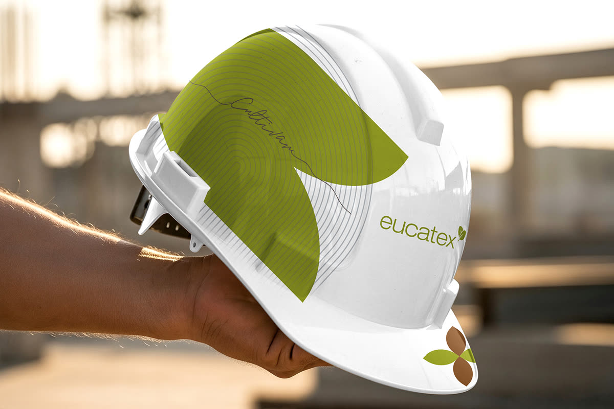 Mão segurando capacete de obra branco com a folha verde e a assinatura Cultivar, com uma obra desfocada ao fundo.