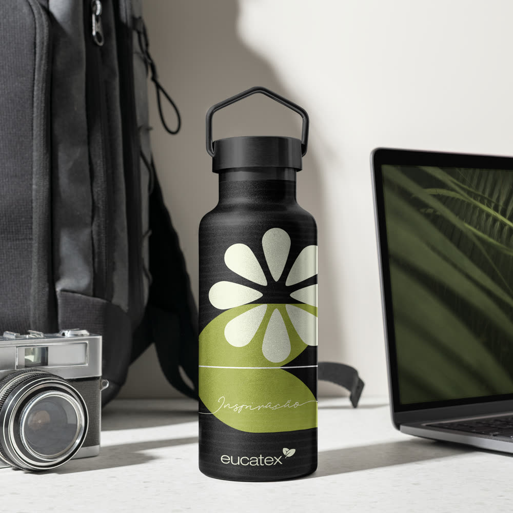Garrafa térmica preta com a flor branca e verde e a assinatura Inspiração, em mesa com mochila, câmera e notebook.