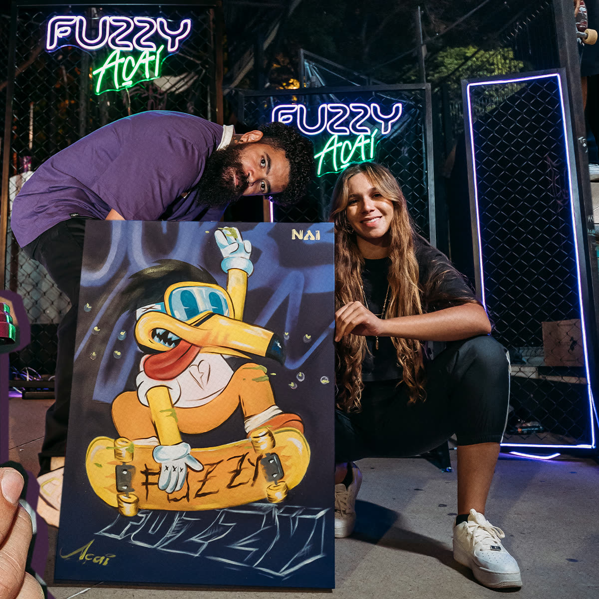 Dupla ao lado do quadro pintado com o mascote do Fuzzy, entre painéis de neon da marca.